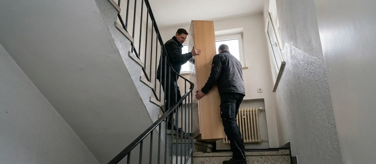 Zwei Träger bugsieren ein hohes schmales Schrankteil um die Wendung eines Treppenhauses